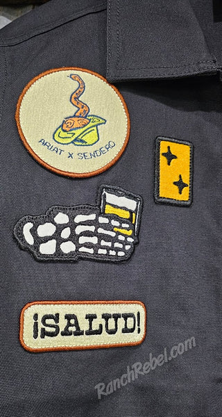 Close-up of a dark shirt with various patches including 'ARIAAT x SEHOGO', a coffee cup, and '¡SALUD!'
